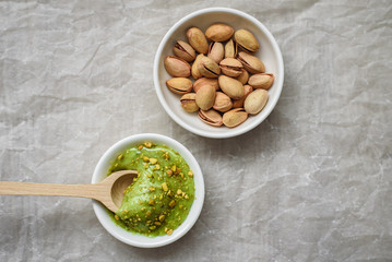 The concept of wholesome food, pistachio paste and unpeeled salted pistachios in white plates, a wooden spoon for pasta on a light background. Components for cooking.