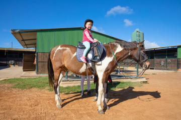 little four years old girl looking at and smiling sitting on a horse outside of stable in a riding center