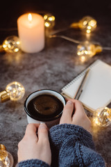 Hand holds  cup of coffee on gray table with notebook, pen and  candle decorated with led lights.Winter and Autumn weekend concept. Flat lay, top view.