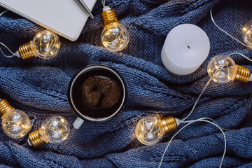  Cup of coffee , candle, notebook, pen and warm woolen sweater , decorated with led lights.Winter and Autumn weekend concept. Flat lay, top view.