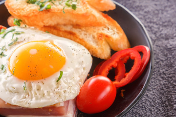 Fried egg with bacon in a black plate with fried pieces of bread, greens and tomatoes on a gray wooden table. Close-up.