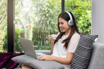 Pretty smiling young woman wear headphones sitting on sofa holding a cup of coffee and usinging on tablet.