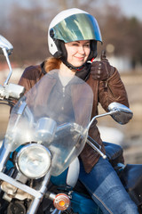 Woman motorcyclist in brown leather jacket and white crash helmet showing thumb up sign with hand