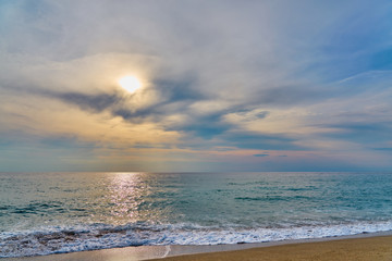                                Sunset at the tropical beach, sun behind clouds reflects on water and waves with foam hitting sand.