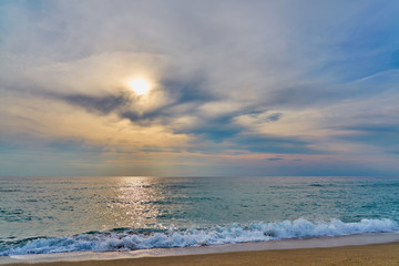                                Sunset at the tropical beach, sun behind clouds reflects on water and waves with foam hitting sand.