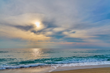                                Sunset at the tropical beach, sun behind clouds reflects on water and waves with foam hitting sand.