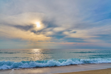 Fototapeta premium Sunset at the tropical beach, sun behind clouds reflects on water and waves with foam hitting sand.