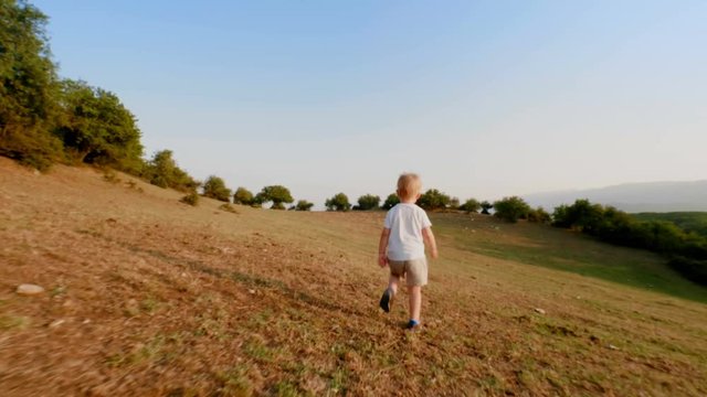 Playful European little boy running along meadow at sunset slow motion steadicam establish shot back view happy child rejoicing at hilly terrain enjoying amazing landscape