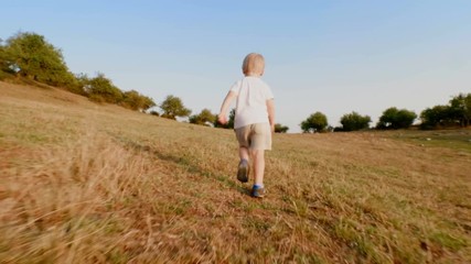 Happy little boy running on meadow with yellow grass in hilly terrain at sunset low angle slow motion steadicam establish shot back view attractive child enjoying walking outdoor
