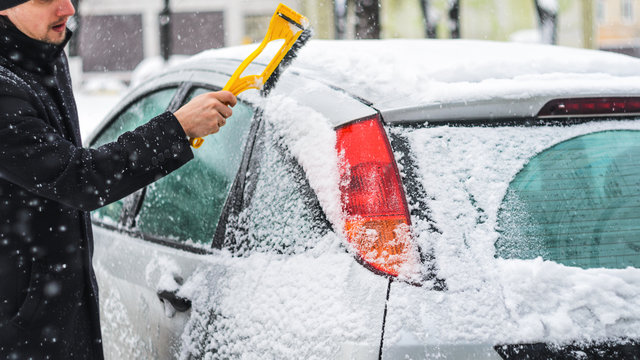 Young man in black coat cleans his car with yellow brush during snowfall. Winter inclement weather. - Powered by Adobe