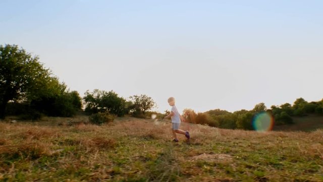 Active baby boy running and enjoying on meadow at sunset slow motion tracking shot European child having good time outdoor at summer evening in middle of beautiful nature side view long shot