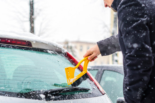 Young man in black coat cleans his car with yellow brush during snowfall. Winter inclement weather.