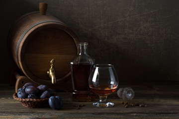 On an old wooden table there is an oak barrel, a bottle, a sniffer with a drink and a wicker dish with plums.