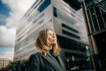 Fototapeta na wymiar Portrait below of young blonde in gray coat on blurred background of city, sky
