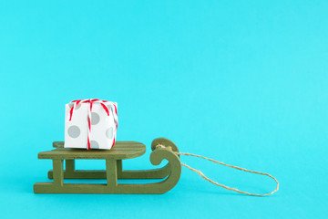 Small gift box wrapped of white and gray paper with red-white ribbon on a green wooden sleigh on a bright blue background. Christmas and New Year concept with copy space.