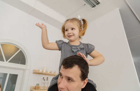 A very beautiful girl is sitting on her dad's shoulders and playing, making faces in the New Year's room.
