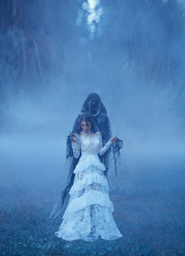 young girl in a white vintage dress and a silver necklace is standing on frozen grass in a thick blue mist in Death captivity and looking down. creative chill color. art photo processing,
