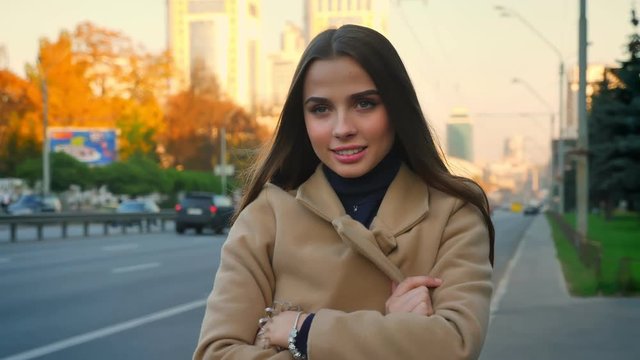 Gorgeous female of caucasian appearance is standing calmly and hidning behing her coat in aoutumn sunny day near road, cars in motion on backgound