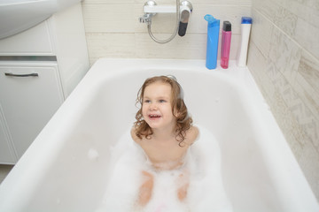 little funny girl in the bathroom playing with foam bath and smiling. Cute baby is washing her hair in bath. The symbol of purity and hygiene education