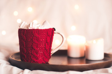 Knitted cup with candle on wooden tray close up over Christmas lights. Winter season.