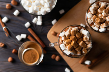 Cups with hot chocolate and marshmallows on cutting board between cinnamon, top view