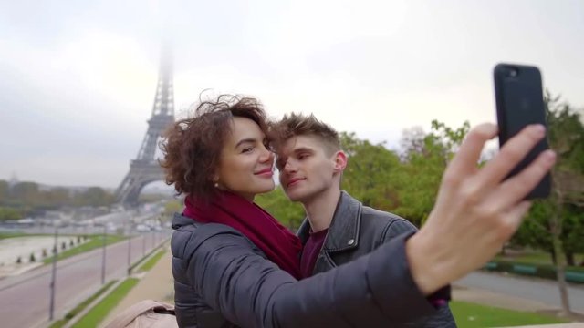 Sweet woman taking selfie of herself and her boyfriend wearing warm jackets in autumn with a Eifferl Tower on a background,
