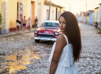 Beautiful dark skinned young lady in white dress standing in front of an old classic car in the old streets of Trinidad in Cuba.
