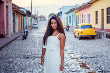 Beautiful dark skinned young lady in white dress standing in front of an old classic car and a motorcycle in the old streets of Trinidad in Cuba.