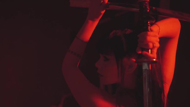 brunette girl stands alone in a scarlet dress from a tank top and skirt, holds a katana weapon in her hands above her head, posing for a perfect photo in red light in the image of a samurai warrior
