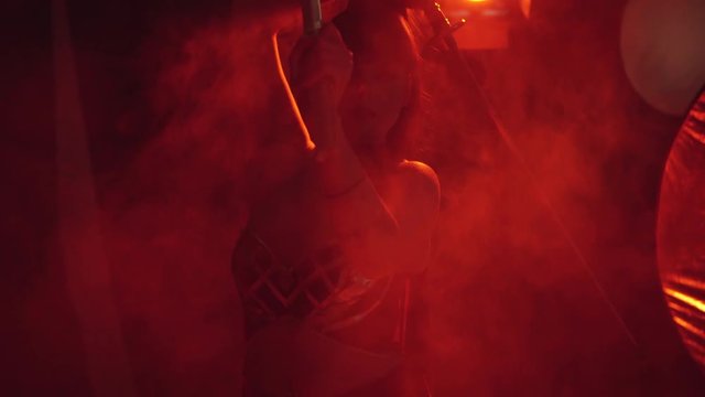 attractive girl with dark hair in red light and fog, smoke, closed her eyes and holding katanas over her head, posing like a samurai warrior, opens her eyes and lowers her sword to the camera