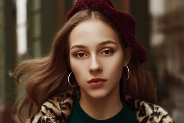 Outdoor close up fashion portrait of young beautiful confident model wearing beret, hoop earrings, leopard print coat, walking in street
