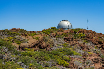Kuppel eines Observatoriums hinter Büschen und Felsen vor einem blauen Himmel