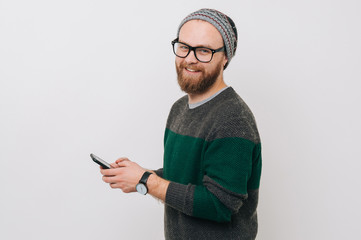Cheerful bearded hipster man using phone over white background and looking at the camera