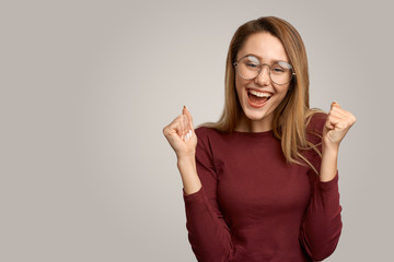A cheerful young woman with positive expression, clenches her fists, and rejoices. Girl with glasses being in high spirits, highlighted on a white background in the Studio. People, happiness, success