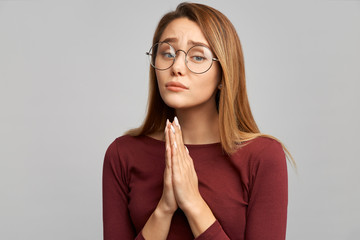 A sweet European woman holds her hands in a prayer gesture, her lips are pressed and her eyes show a request, begging for something. Isolated on white background in Studio