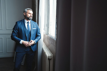 Thinking about... Thoughtful man in full suit looking away through the window while standing indoors.