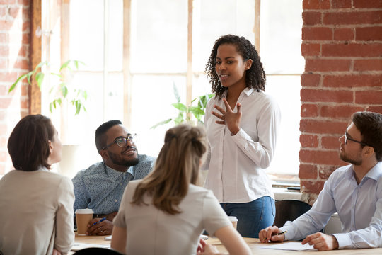 African American woman standing and talking about new project, opinion, business strategy at briefing, company meeting, multiracial colleagues group looking attentively at speaking businesswoman