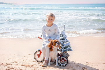 Santa claus kid in Christmas dress. Happy child on the bicycle or tricycle near water with a dog. Xmas party celebration, childhood. Winter holiday vacation. New year small boy at sea beach.
