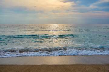Fototapeta premium Sunset at the tropical beach, sun behind clouds reflects on water and waves with foam hitting sand.