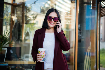 Portrait pretty girl with dark hair in Burgundy fur coat of artificial fur. She looks excited and talking on a mobile phone. City lifestyle.