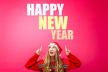 Beautiful girl in a Santa hat, with a tinsel around her neck, pointing fingers up and showing a blank space with text. Happy New Year. on a red background.