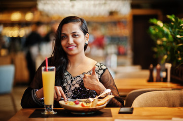Pretty indian girl in black saree dress posed at restaurant, sitting at table with juice and salad. Show thumb up.