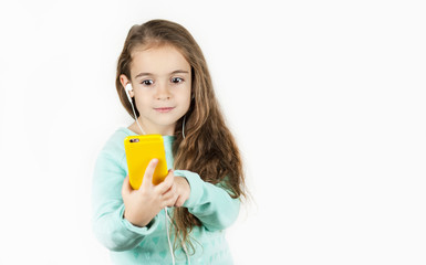 Bright beautiful little girl in headphones with the phone emotionally posing for a selfie on an isolated background.