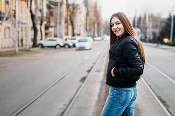 Portrait from back of elegant girl with long hair walking on city background. She has black leather jacket and jeans. She is smiling camera.