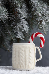 Obraz na płótnie Canvas Knitted winter cup of hot chocolate with candy cane on snowy table under christmas tree