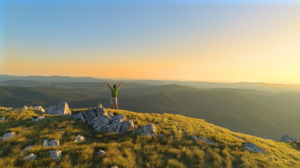 AERIAL: Young man runs up to top of the hill and victoriously raises his arms.
