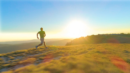 LENS FLARE: Athletic young man sprints down a grassy hill on a sunny morning - obrazy, fototapety, plakaty