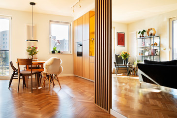 Cozy scandinavian open space with dining, living room and kitchen with design sofa and family table. Bright and sunny room with big windows and brown wooden parquet. 