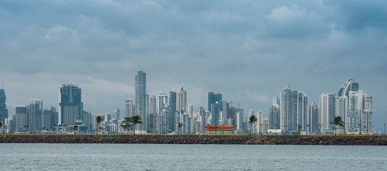 Hot, humid day in Panama city as another rainstorm brews quickly over the city skyline.  Tall buildings shimmer in heatwaves rising in humid air.  People on Panama Canal jetty park in foreground.