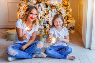 Happy family woman mother and little girl relax playing sparkler near Christmas tree on Christmas eve at home. Mom, daughter in house with winter decoration. Christmas New Year time for celebration